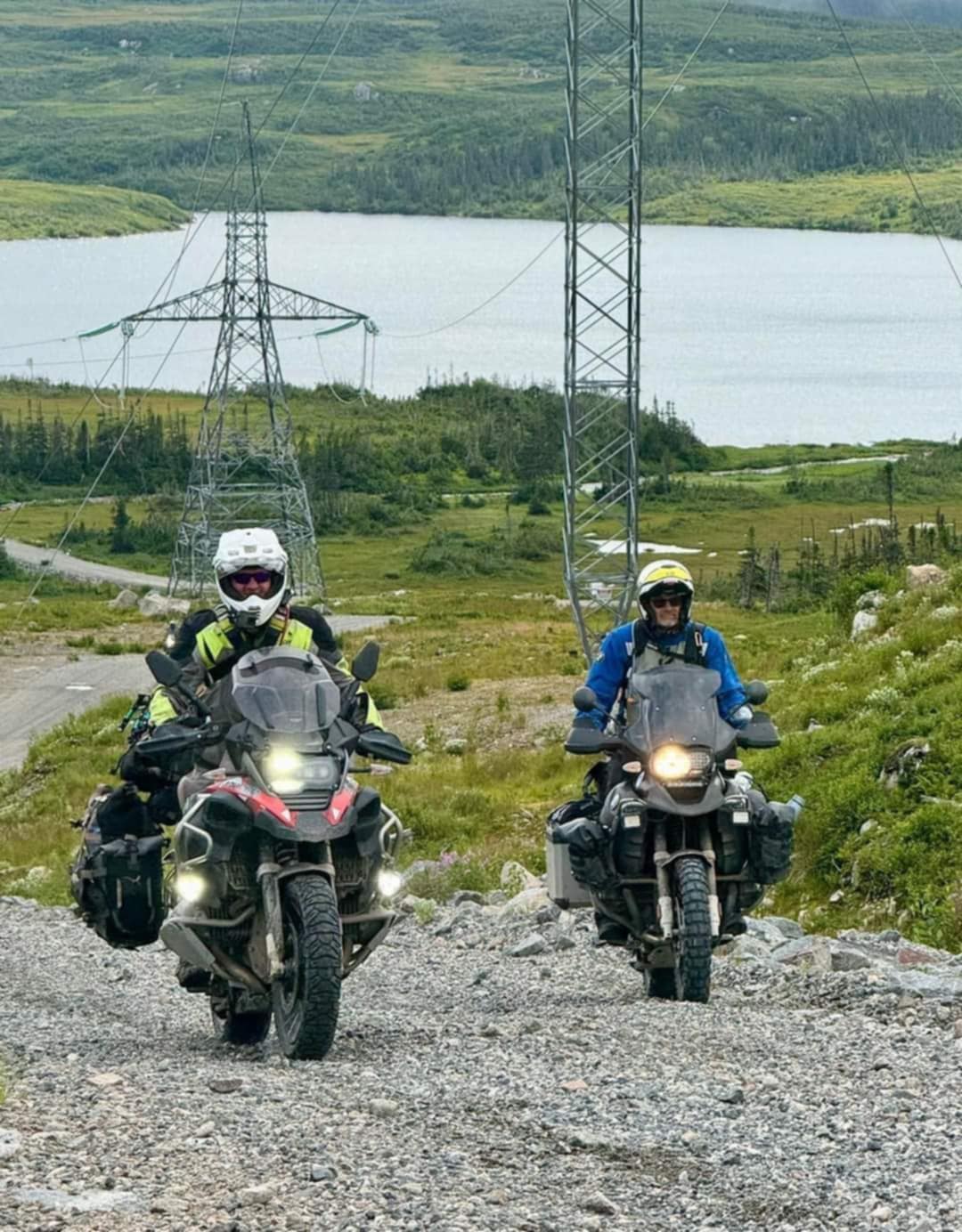 two adventure bike riders coming up a hill with power lines and water in the background